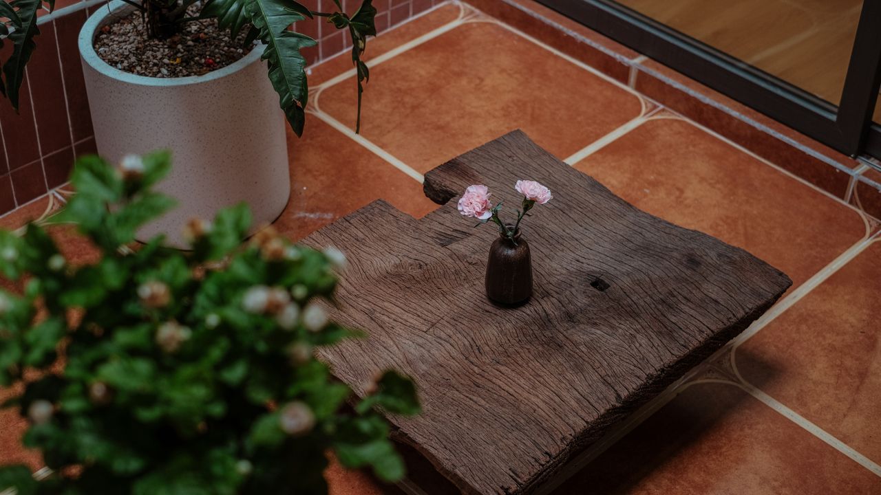 A rustic wooden table with pink flowers in a small vase, surrounded by plants.